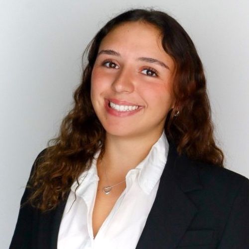 Smiling woman with long brown hair wearing a black blazer and white shirt against a plain background.