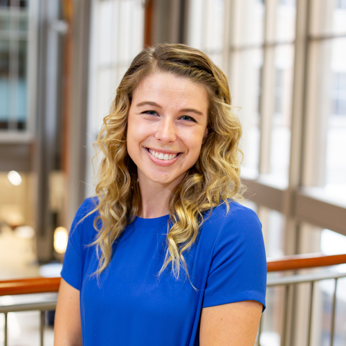 Tess Watt Headshot in front of wall of windows wearing royal blue with blonde curly hair