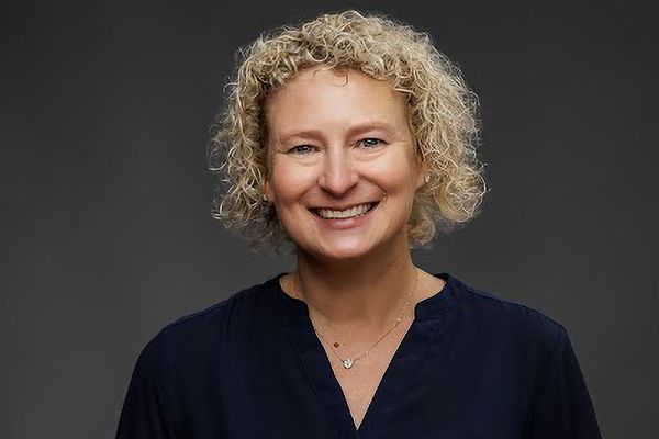 Smiling person with curly blonde hair wearing a dark blouse against a dark gray background.
