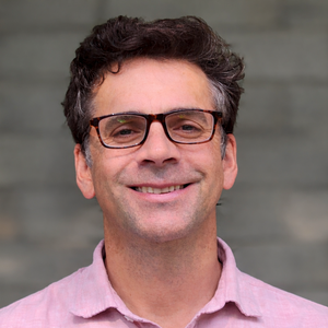 A smiling person wearing glasses and a pink shirt is standing in front of a gray brick wall.