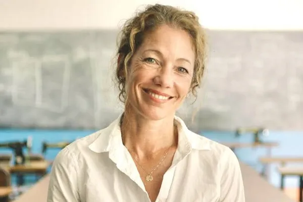 Smiling woman with curly hair in a white shirt stands in a classroom with sewing machines and a chalkboard in the background.