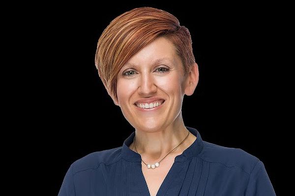 Smiling person with short, styled hair, wearing a dark blue top and a necklace, against a black background.