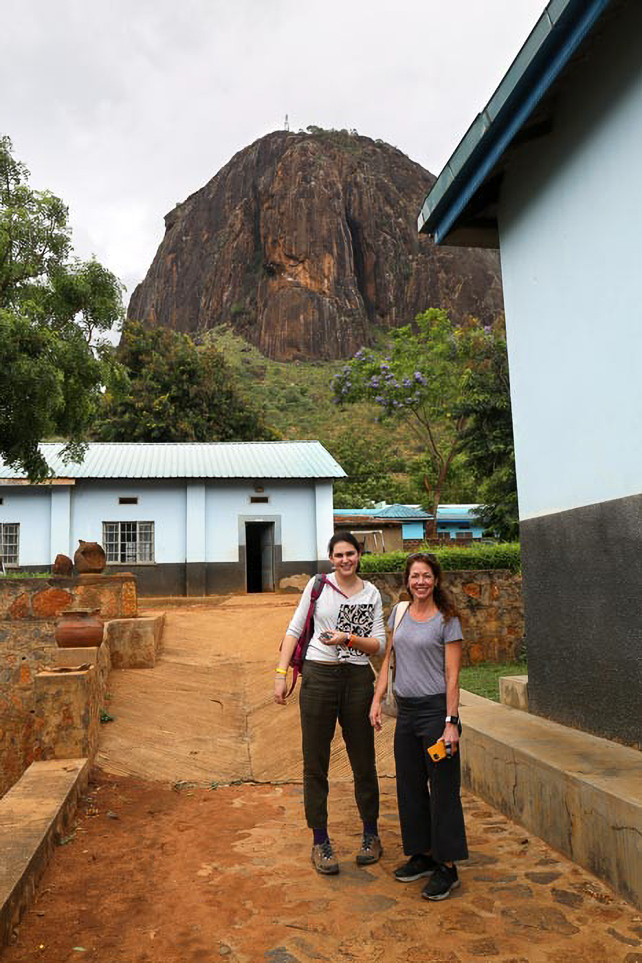 Two people stand smiling in front of a building with a large rocky hill in the background, under a cloudy sky.