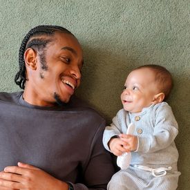 A father lies on the ground smiling at his baby, who smiles back.