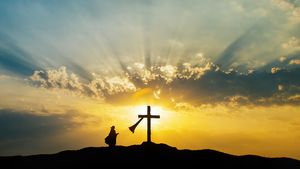 Silhouette of a person kneeling beside a hilltop cross as golden sunrise rays burst through clouds.