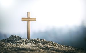 Wooden cross planted in a mound of earth with a misty, blurred background and bright sky.