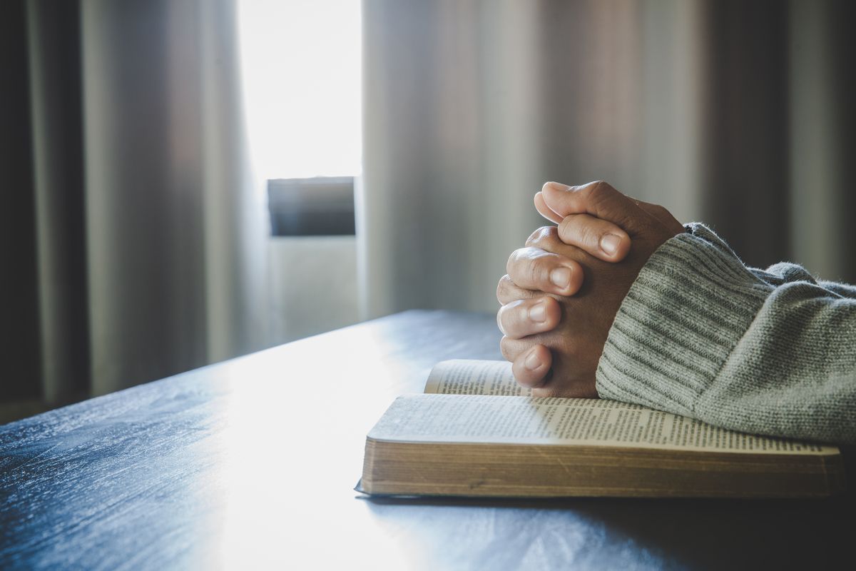 Hands clasped in prayer rest on an open book on a table, with soft window light and curtains in the background.