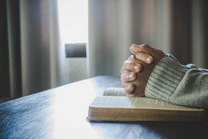 Hands clasped in prayer rest on an open book on a table, with soft window light and curtains in the background.
