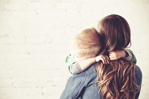 Woman with long hair in denim jacket holds toddler asleep on her shoulder, child's arms wrapped around her neck, white brick background.