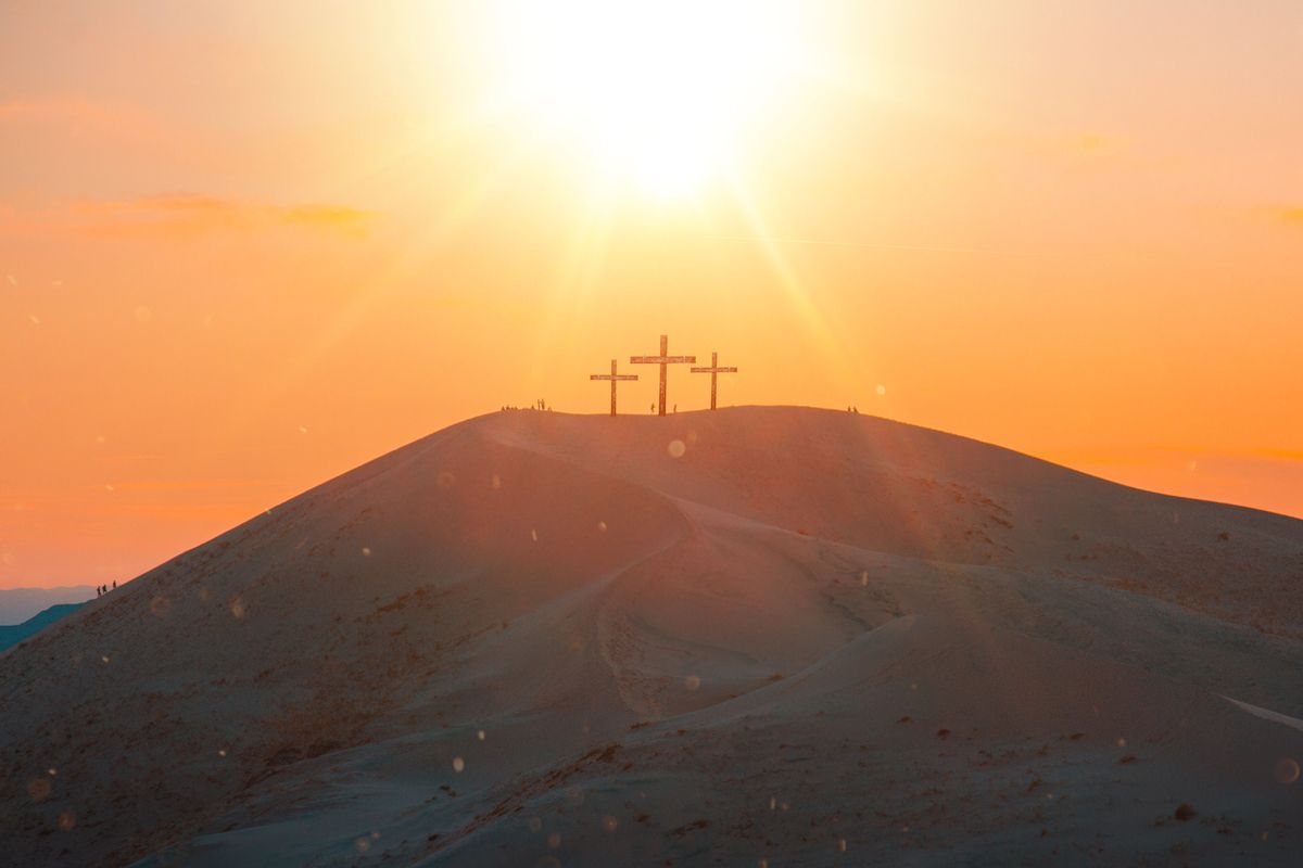 Three wooden crosses atop a sandy hill at sunset, sunbeams radiating over dunes with tiny silhouetted figures.