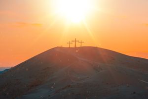 Three wooden crosses atop a sandy hill at sunset, sunbeams radiating over dunes with tiny silhouetted figures.