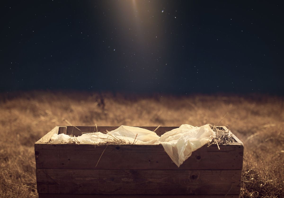 Wooden creche filled with straw and a white cloth in a grassy field at night under a starry sky.