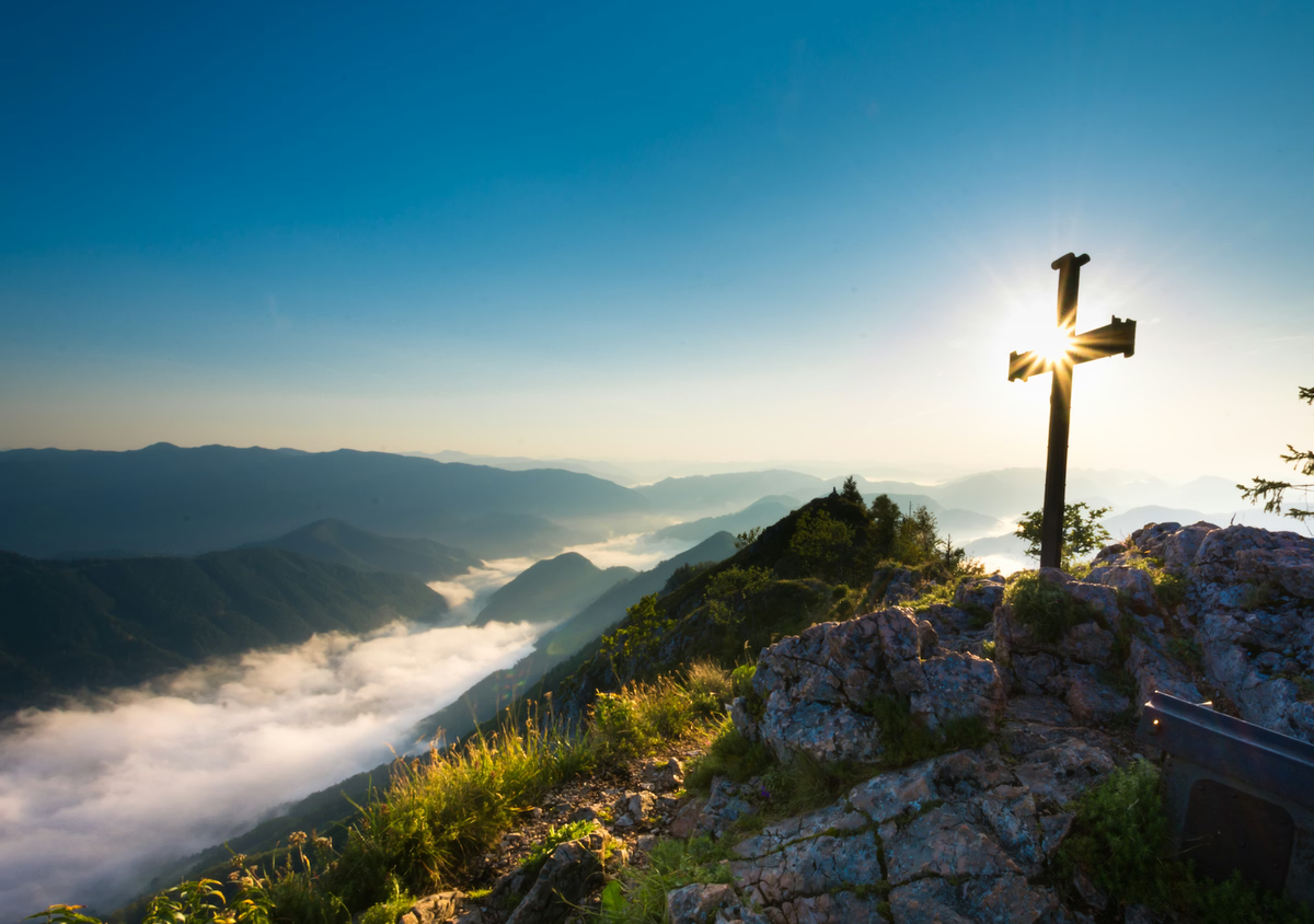 The sun shines through a cross on a mountaintop.