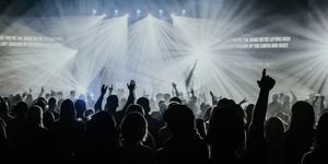A group of people in a rock-concert-style worship service with bright lights obscuring the people on stage and many silhouetted people with hands raised.
