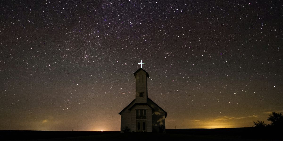 A church is photographed against a clear, starry sky. The cross on top of the steeple is brightly illuminated and there are glowing lights illuminating wispy clouds just over the horizon.