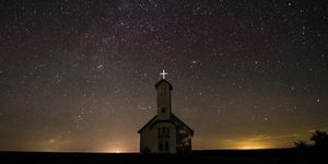 A church is photographed against a clear, starry sky. The cross on top of the steeple is brightly illuminated and there are glowing lights illuminating wispy clouds just over the horizon.