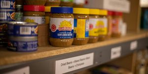 A shelf of food in a food pantry with tuna fish in cans in the foreground and sunflower butter in a jar at the focal point.