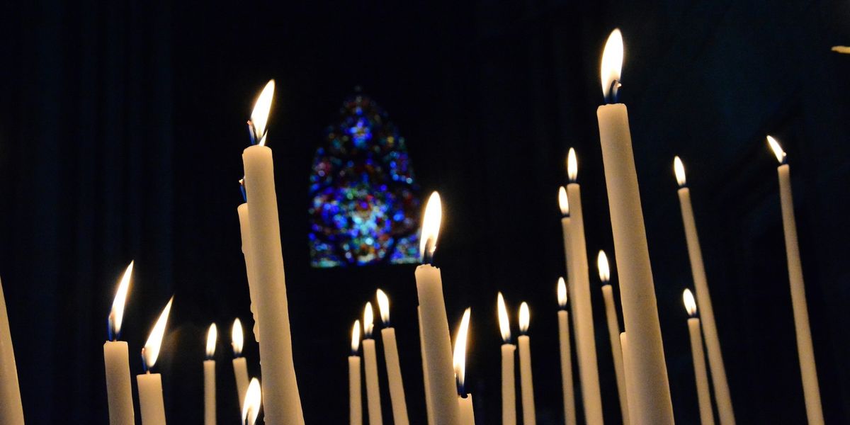 Tall white candles with flickering flames in a dark setting, stained glass window softly illuminated in the background.