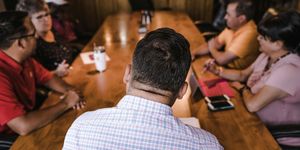 Depicts the back of a middle-aged man's head and shoulders. Before him, out of focus, is a table with four people at it, who appear to be having a discussion.