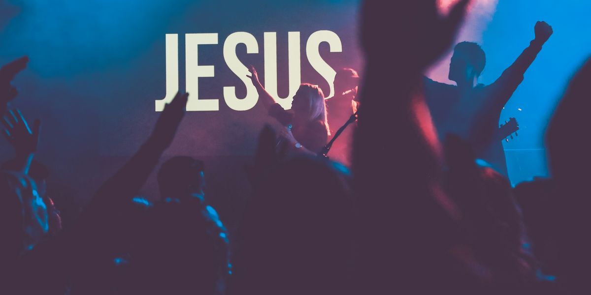 A group of people on stage singing in front of a brightly lit sign saying "JESUS." A blond woman in the center holds a microphone with her hand in the air along with another musician. They are in a dark room and can be seen through the silhouettes of the audience's raised hands.