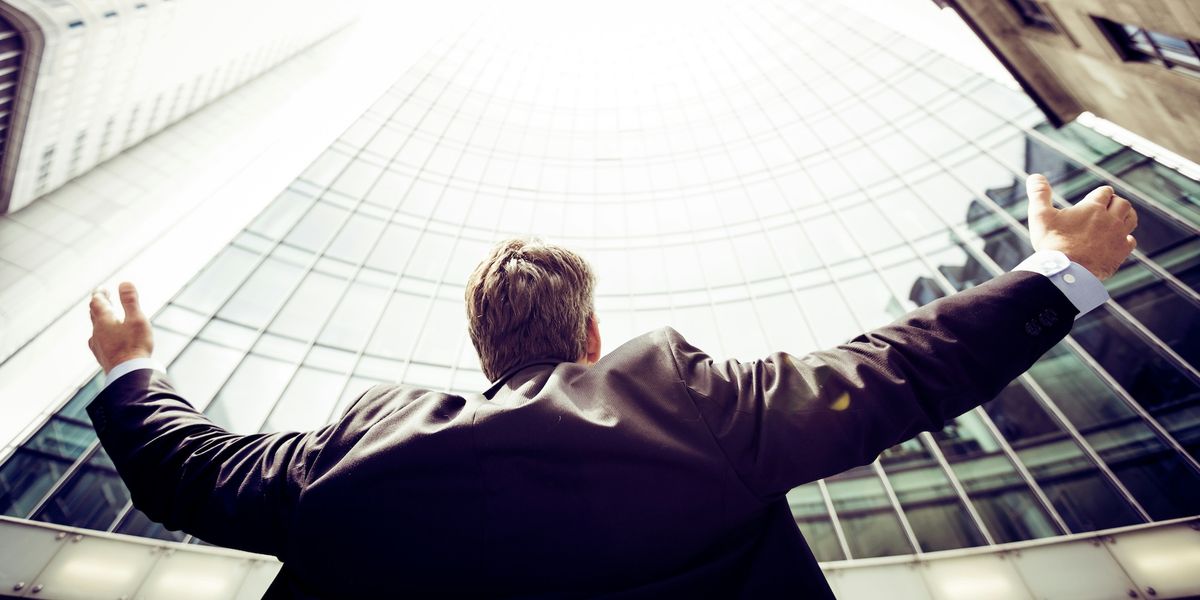A man in a suit looks up at a wall of windows on a skyscraper from below, with his arms spread out.
