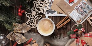 A cup of coffee or cocoa on a table surrounded by festive clutter including spruce boughs, ribbons, walnuts, candles, glass ornaments, cut-out wooden ornaments, holly berries, cinnamon sticks, and envelopes.