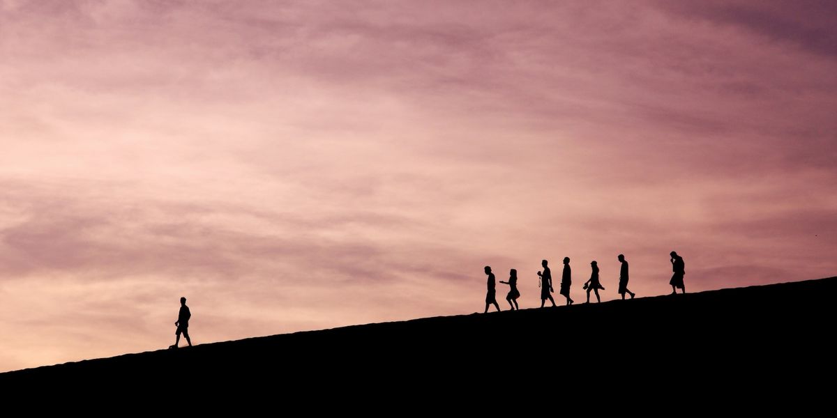 A line of silhouetted figures against sunset clouds descending down a slight slope. One figure is far ahead of the group of others.