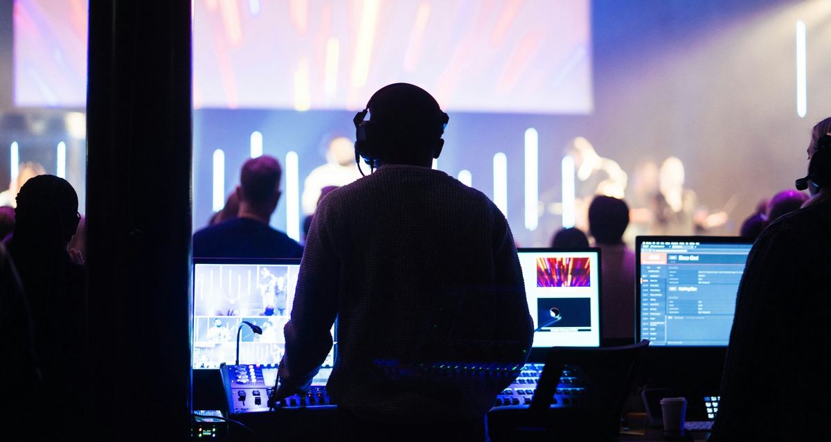 A person faces screens and sound boards while a band plays in a dark room.