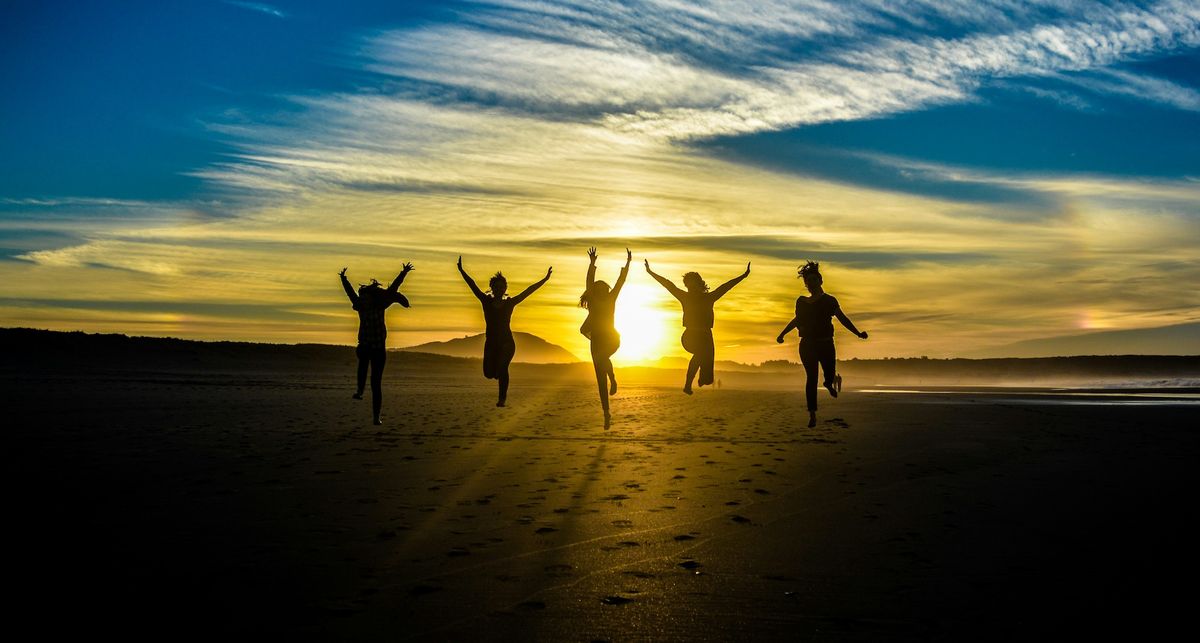 Five people jumping at once on a sandy beach, silhouetted against a rising sun. The sky has angular high clouds with a faint rainbow showing on either side of the sun.