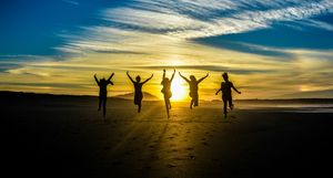 Five people jumping at once on a sandy beach, silhouetted against a rising sun. The sky has angular high clouds with a faint rainbow showing on either side of the sun.