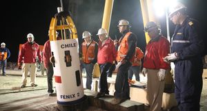 A cylindrical metal frame sits on the ground, affixed to a cable at the top. The bottom three quarters is covered in thin metal sheeting and has small wheels oriented vertically. It is labeled Chile and Fenix two. A line of individuals in hard hats and high visibility gear are looking at it.