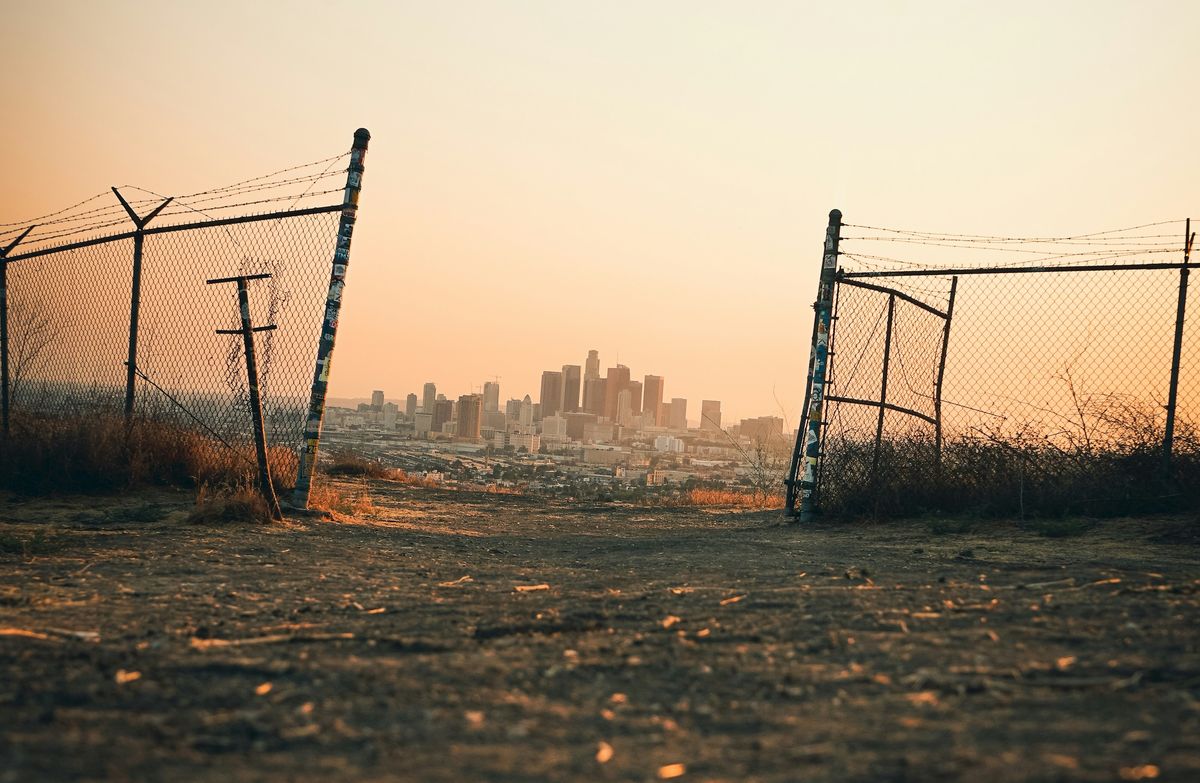 An open barbed-wire fence leads to a city.