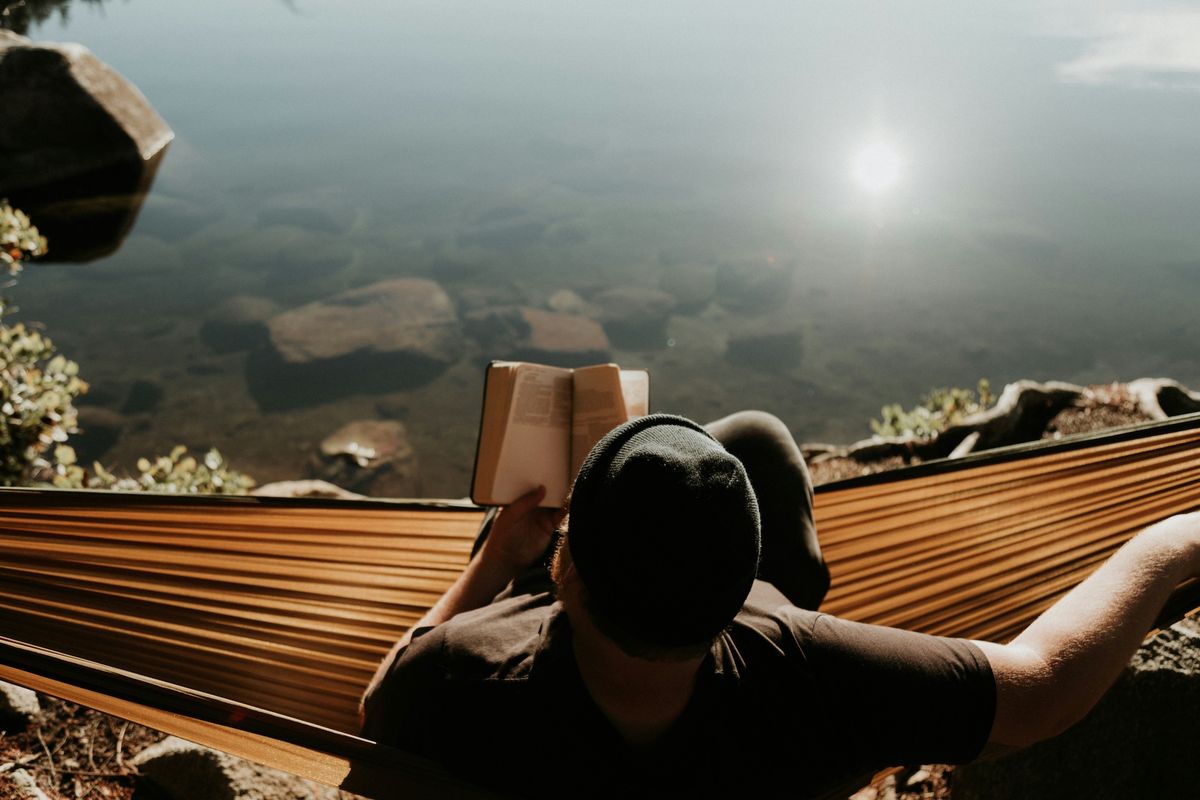 A man reads a Bible in a hammock by a lake.