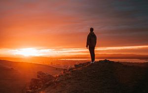 A man stands on top of a hill and watches the sun rise over distant ridges.