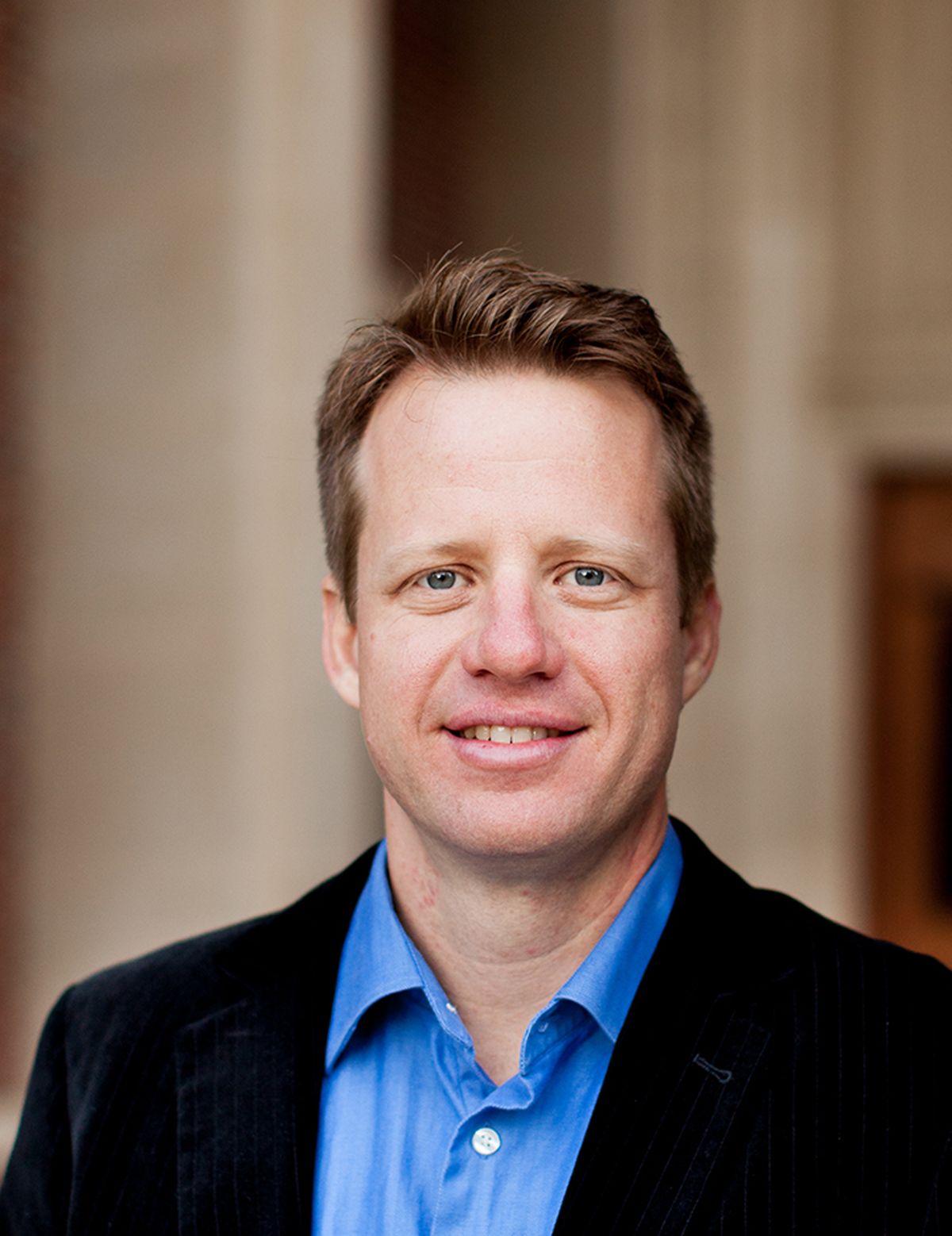 A white man with brown hair wearing a jacket and blue shirt.