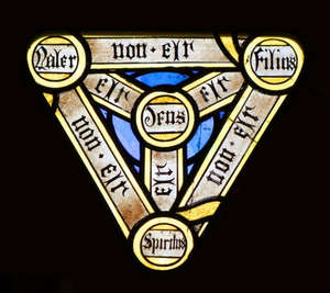 Triangular medieval stained-glass panel with three corner medallions labeled "Father, "Son" and the "Holy Spirit" in Latin.