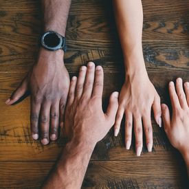 Four hands with different skin tones on a dark brown wood surface.