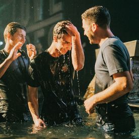 A newly baptized man drips with water standing in a pool with two other men.