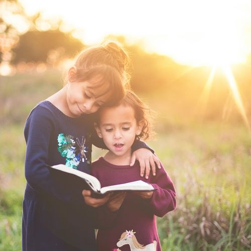 Two young girls stand together reading a bible in a meadow.