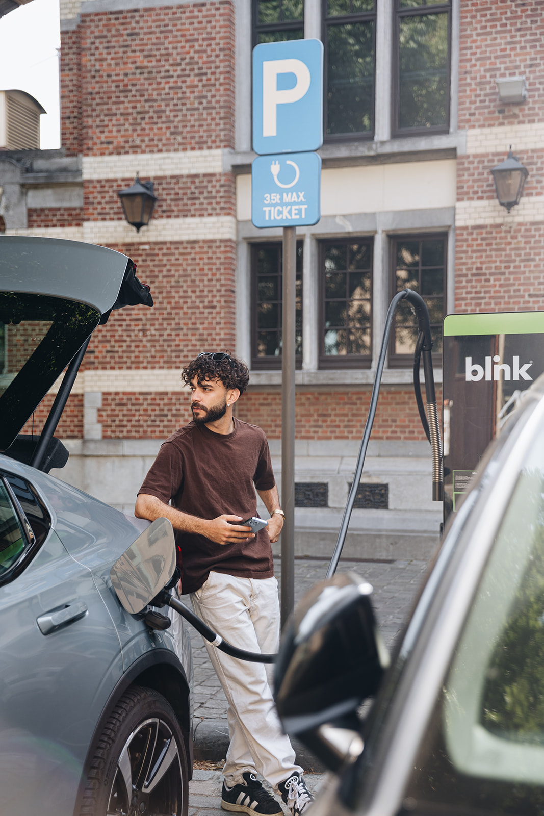 Man in casual clothing stands by a charging electric car, holding his phone. Brick building and parking sign visible in the background.