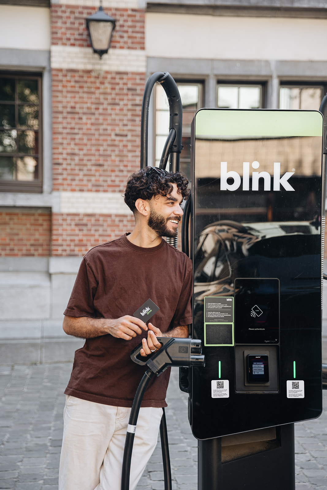 Man charging electric vehicle at a Blink charging station, holding a card. Brick building in the background.