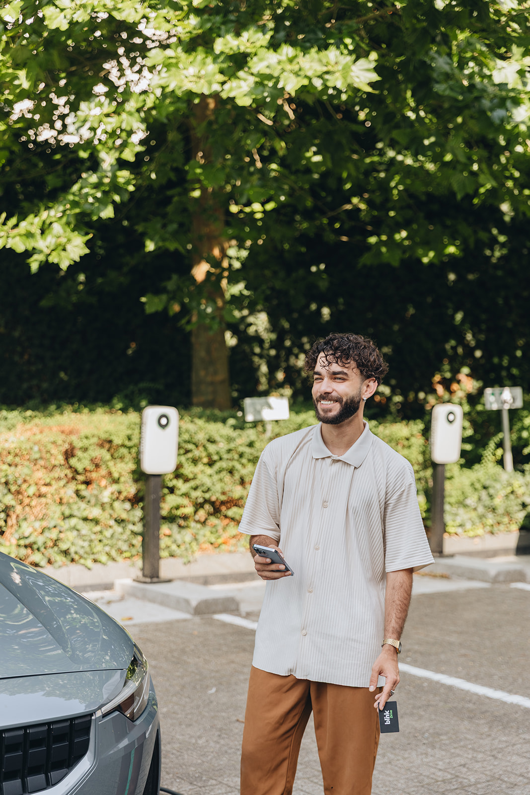 Man stands smiling near an electric car, holding a phone and key. Green foliage and charging stations in the background.