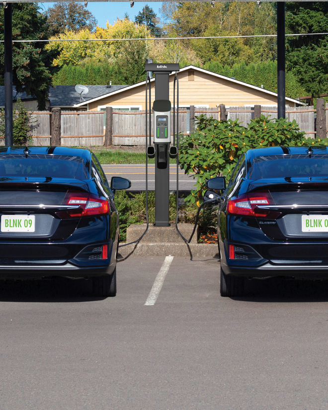 Two black electric cars parked at charging stations labeled "Blink 09" in a sunny parking lot, with trees and a fence in the background.
