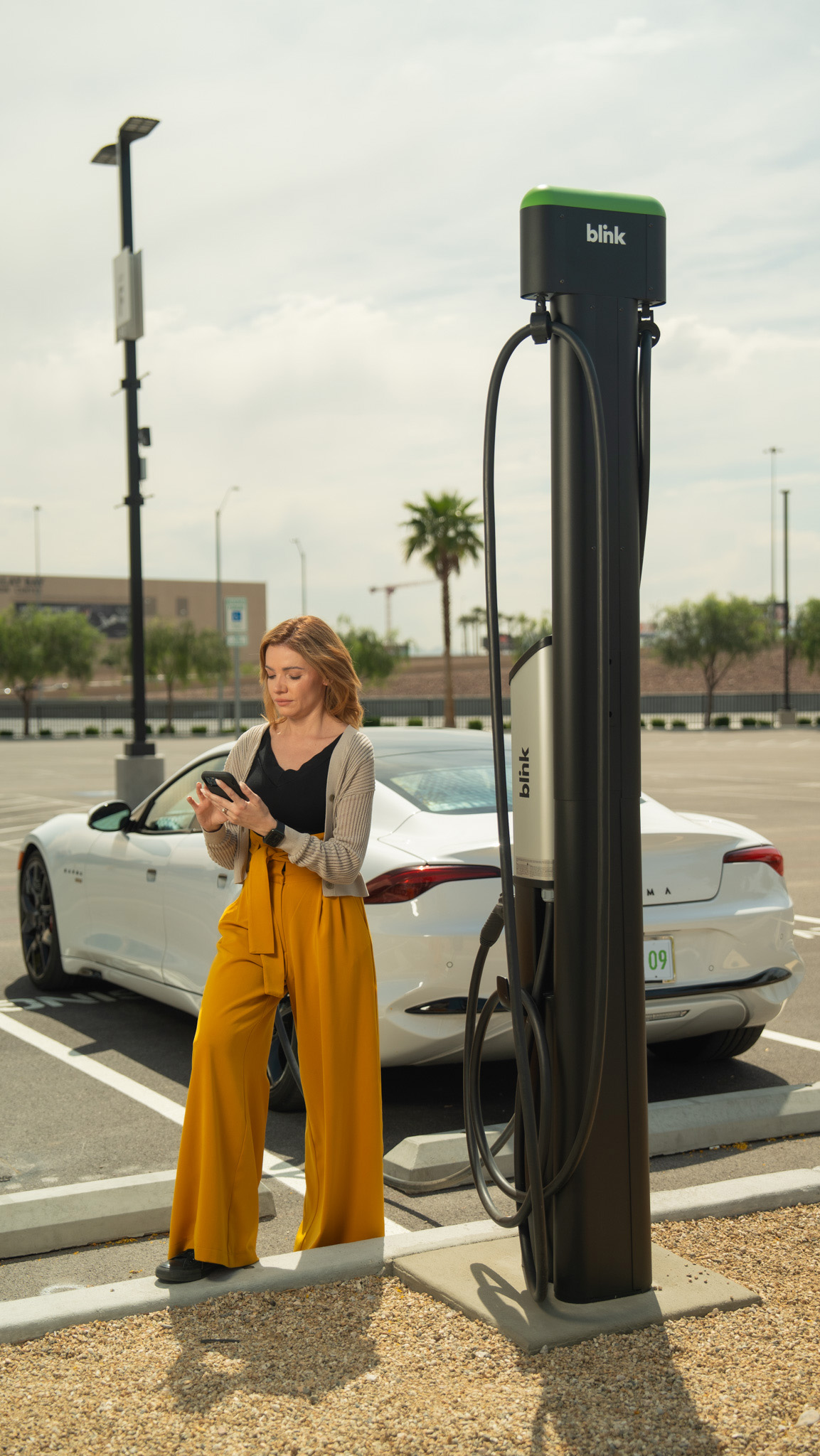 A woman stands by an electric vehicle charging station, using her phone. A white car is parked nearby on a sunny day.