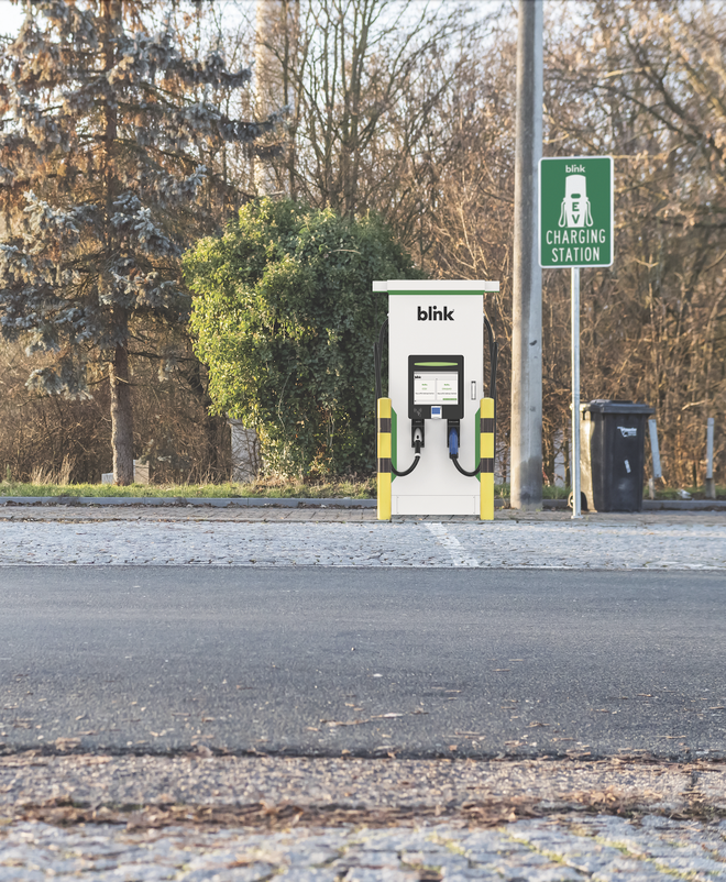 Electric vehicle charging station labeled "blink" beside a road, with trees and a charging station sign in the background.