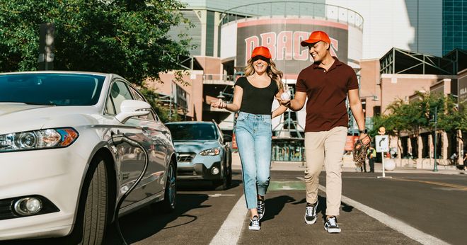 A smiling couple wearing red caps walks hand-in-hand across a city street in daylight, with cars and a building in the background.