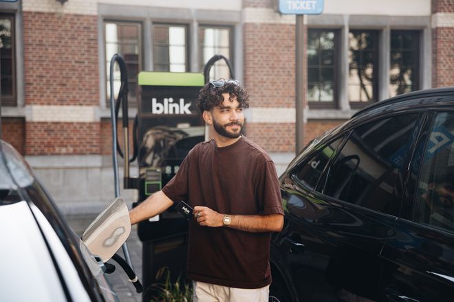 Man with curly hair charging an electric car at a Blink station, holding a card and smiling. Brick building in the background.