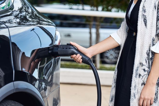 Person charging an electric car outdoors, holding the charger plug connected to the vehicle. Trees and another car are visible in the background.