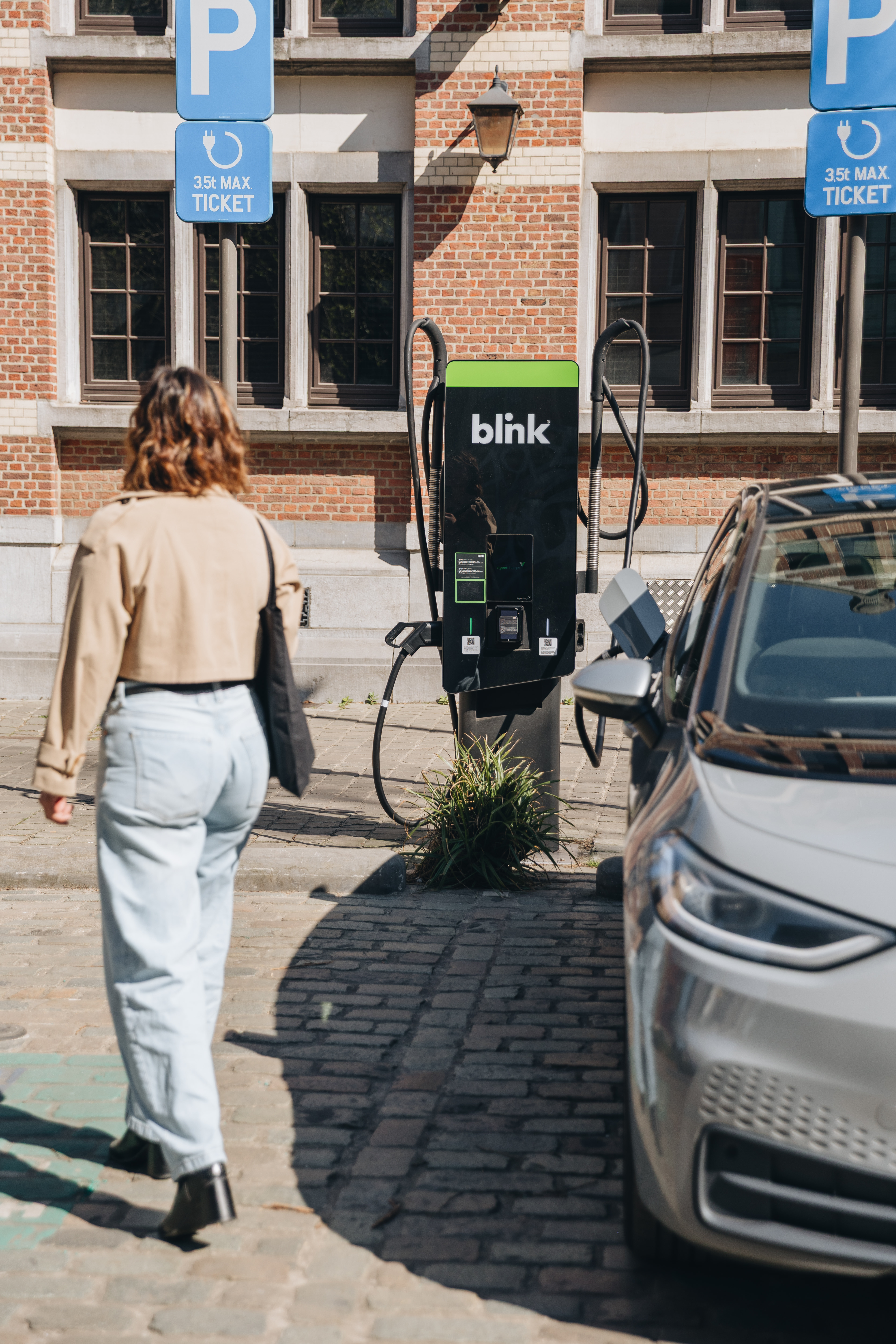A woman walks past a parked electric car charging at a Blink station on a cobblestone street with building and parking signs in the background.