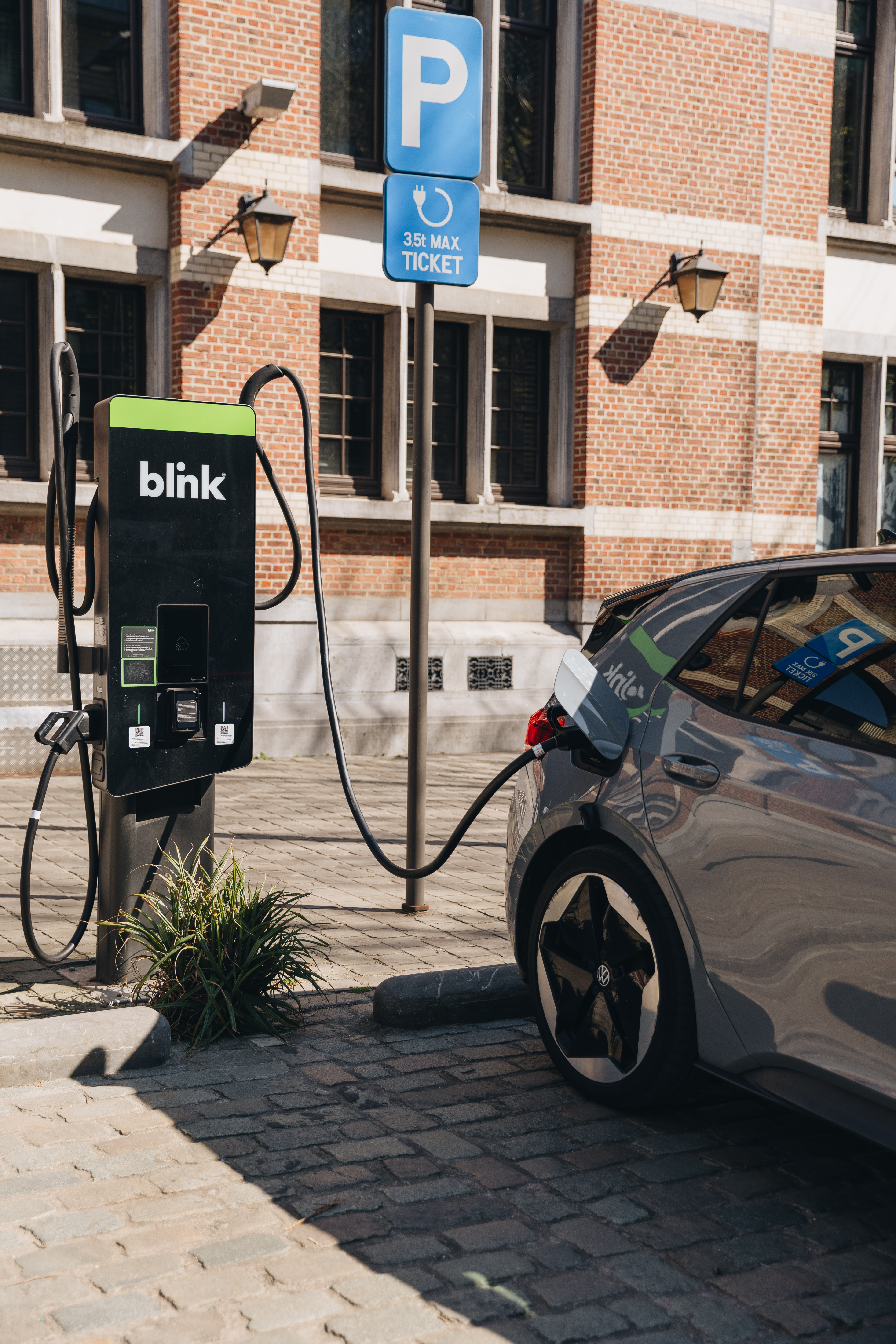 Electric car charging at a Blink station in a cobblestone parking area, near a brick building. A parking sign reads "35€ Max Ticket."